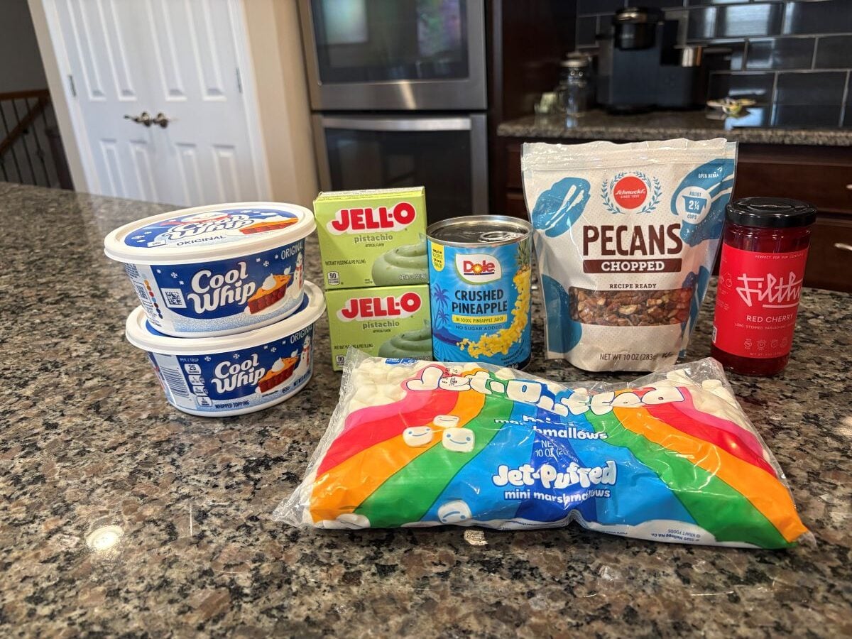 Assorted dessert ingredients for a classic Watergate Salad recipe: two tubs of Cool Whip, two boxes of pistachio Jell-O, crushed pineapple, chopped pecans, red cherries, and mini marshmallows on a kitchen counter.