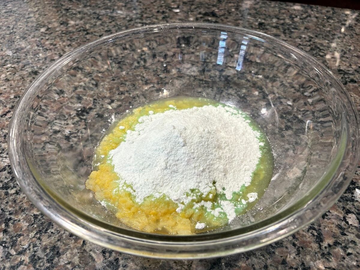 A glass bowl on a granite countertop contains a pile of white flour atop a mixture of mashed banana and egg&mdash;perfect for pairing with a pistachio salad recipe or classic Watergate Salad for your next gathering.