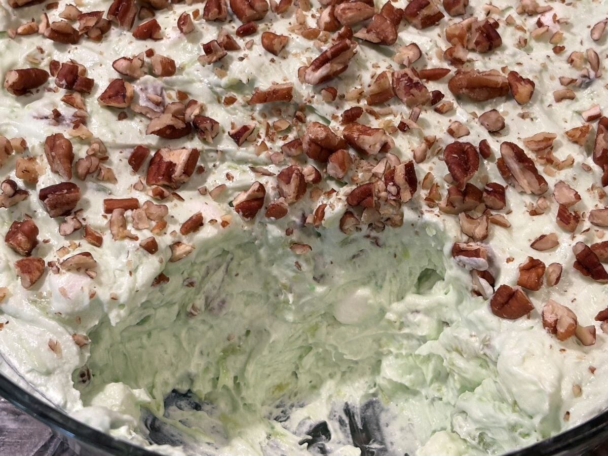 A close-up of a creamy, light green pistachio salad topped with chopped pecans, with a portion scooped out to reveal the fluffy texture inside.