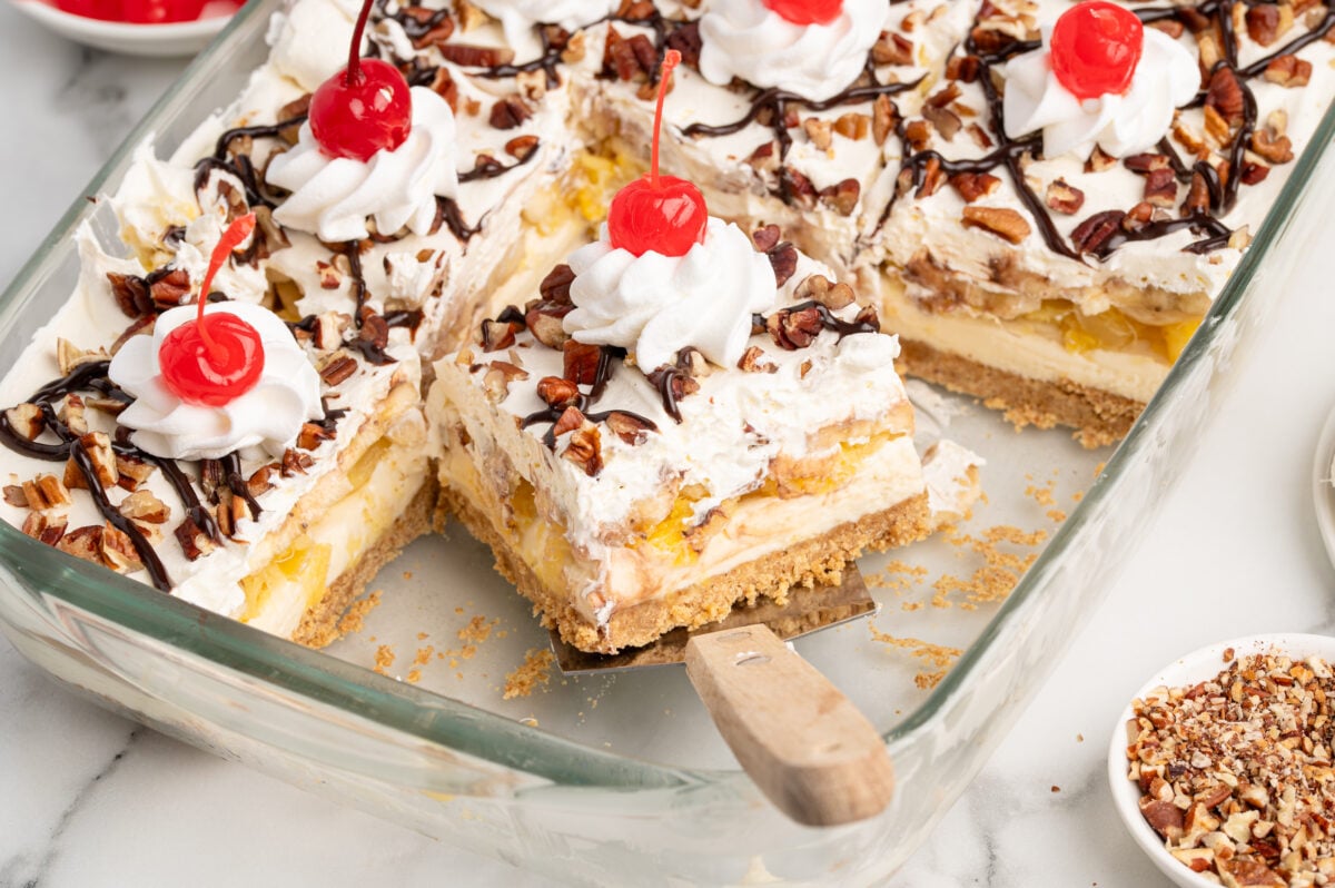 A glass baking dish with slices of Banana Split Cake topped with whipped cream, cherries, chopped nuts, and chocolate drizzle; a spatula lifts one slice. Small bowls of cherries and nuts complete this tempting Banana Split Cake Recipe.