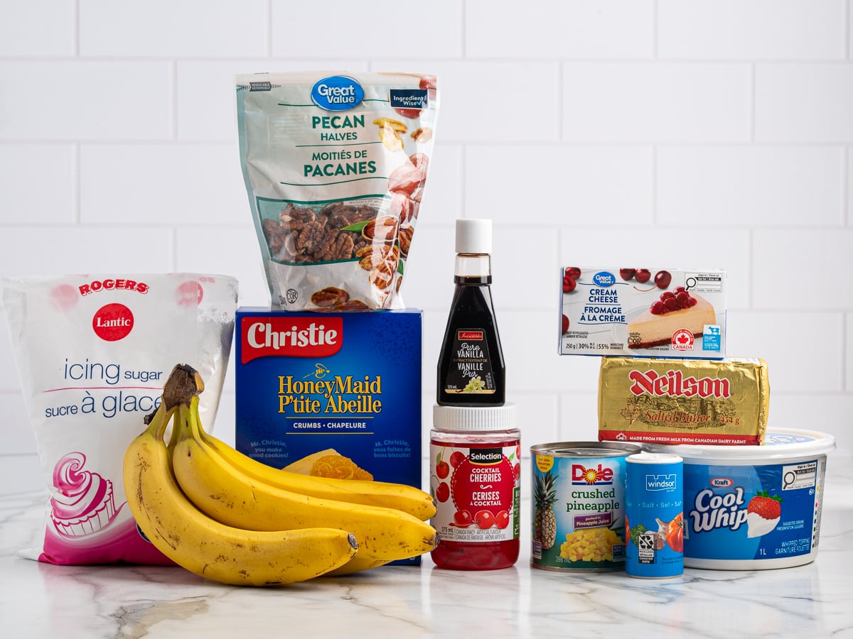 A variety of baking ingredients on a counter, perfect for making a Banana Split Cake, including bananas, icing sugar, graham crackers, pecan halves, vanilla, whipped topping, butter, canned pineapple, pie filling and cream cheese.