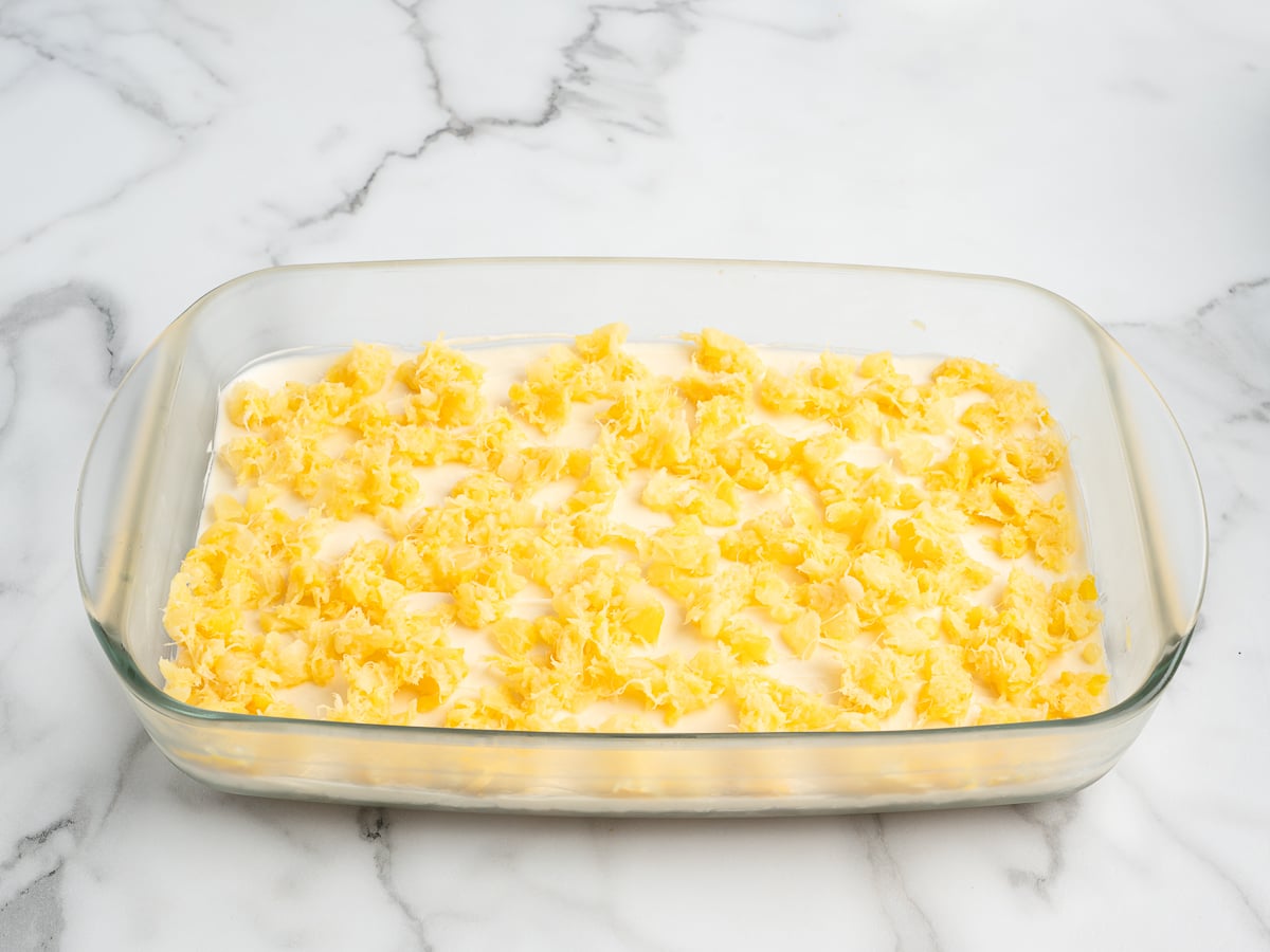 A clear glass baking dish filled with an uncooked Banana Split Cake mixture, topped with small, uneven dollops of yellow dough or butter, set on a white marble surface.