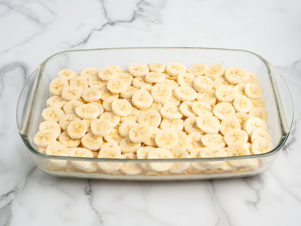 A glass baking dish filled with evenly layered banana slices, perfect for preparing a classic Banana Split Cake, sits on a white marble surface.
