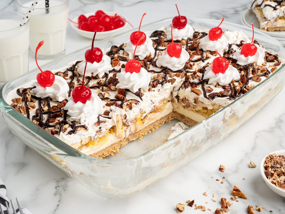 A glass baking dish holds a Banana Split Cake layered dessert, topped with whipped cream, cherries, nuts, and chocolate drizzle. Slices are missing. Plates, milk glasses, and bowls of cherries and nuts surround it on a marble surface.