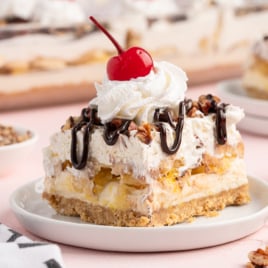 A slice of Banana Split Cake with a graham cracker crust, whipped cream, nuts, chocolate drizzle, and a cherry on top is served on a white plate—try this Banana Split Cake Recipe for the ultimate classic dessert experience.