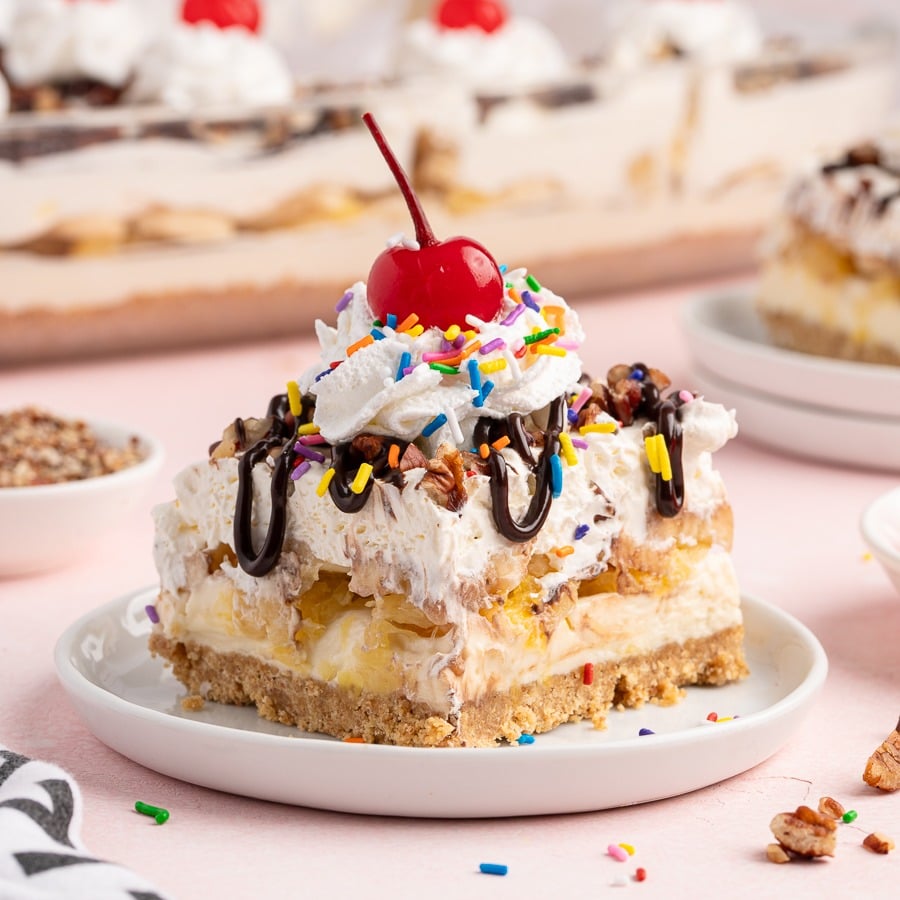 A slice of Banana Split Cake is served on a white plate, topped with whipped cream, chocolate syrup, colorful sprinkles, chopped nuts, and a cherry. This Banana Split Cake Recipe features layers of banana, cream, and a crumbly crust.