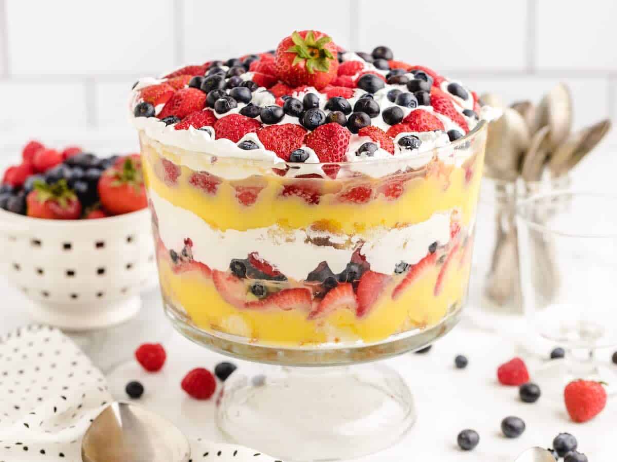 A glass trifle bowl filled with layers of cake, yellow custard, whipped cream, and assorted berries, topped with strawberries and blueberries showcases this classic Vanilla Trifle Recipe. A bowl of berries and utensils are in the background.