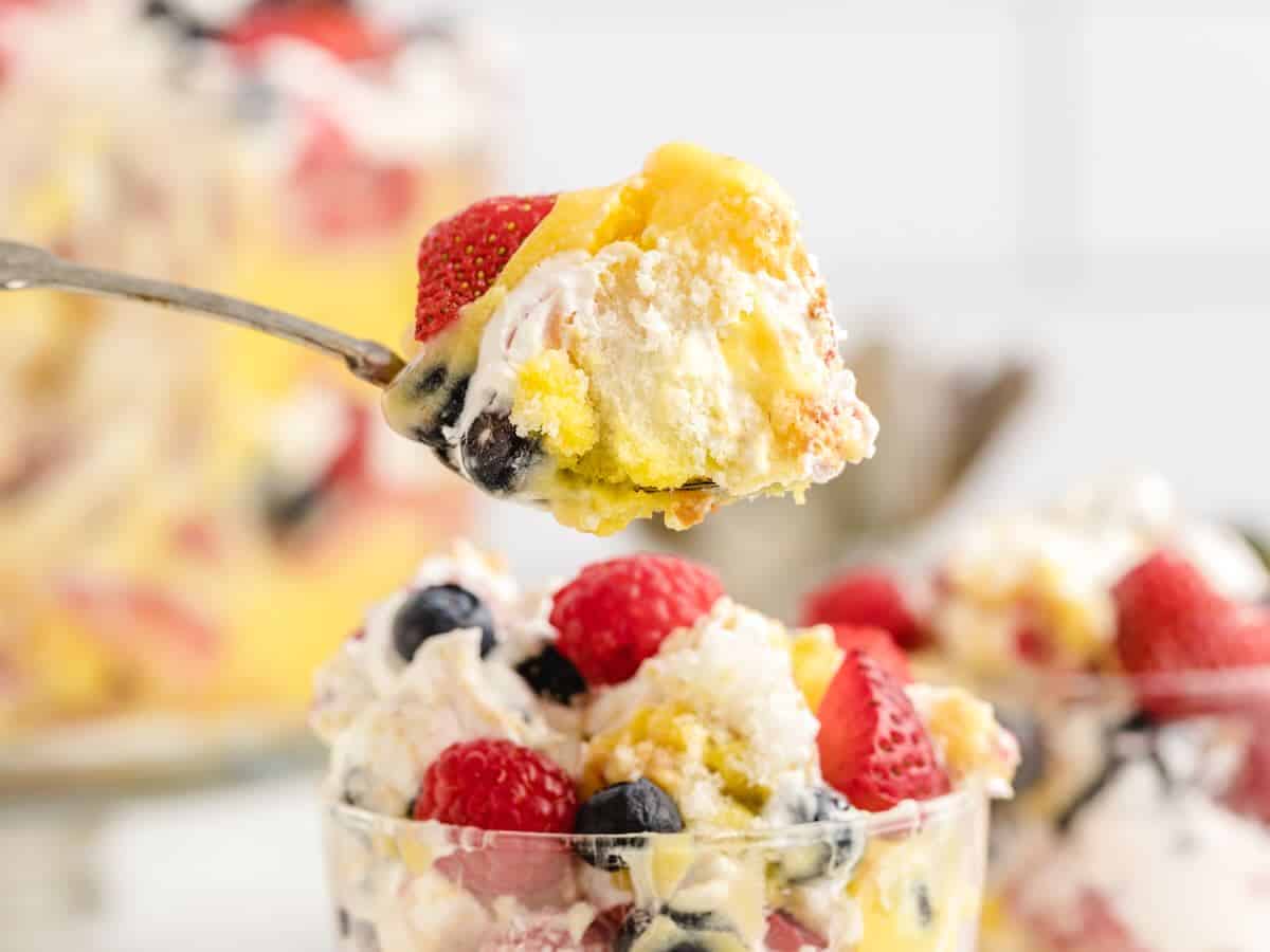 A close-up of a spoonful of berry trifle with whipped cream, strawberries, blueberries, cake, and custard—perfect for anyone seeking a classic Vanilla Trifle Recipe. More trifle is seen in a glass bowl below, with a beautifully blurred background.