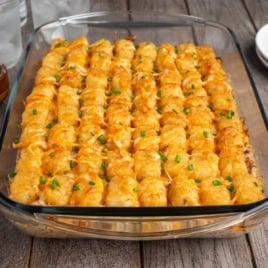 A glass baking dish filled with Cowboy Casserole, topped with melted cheese and chopped green onions, sits on a wooden table. This hearty Cowboy Casserole recipe is sure to be a crowd-pleaser at any meal.