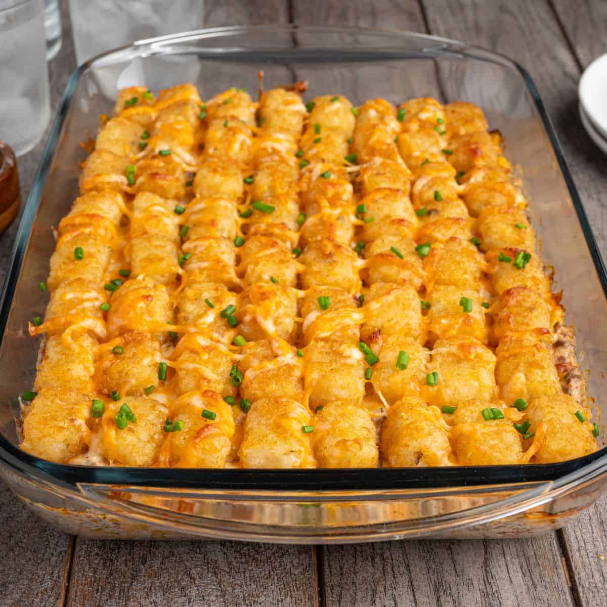 A glass baking dish filled with Cowboy Casserole, topped with melted cheese and chopped green onions, sits on a wooden table. This hearty Cowboy Casserole recipe is sure to be a crowd-pleaser at any meal.