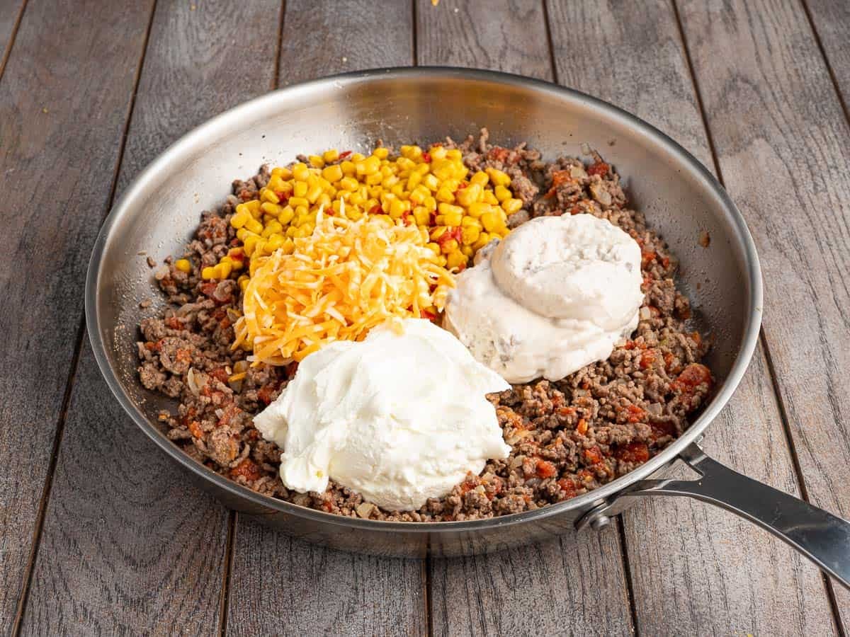 Cooked ground beef in a skillet on a wood surface, with corn, shredded cheese, sour cream and cream of mushroom soup added and not yet stirred in.