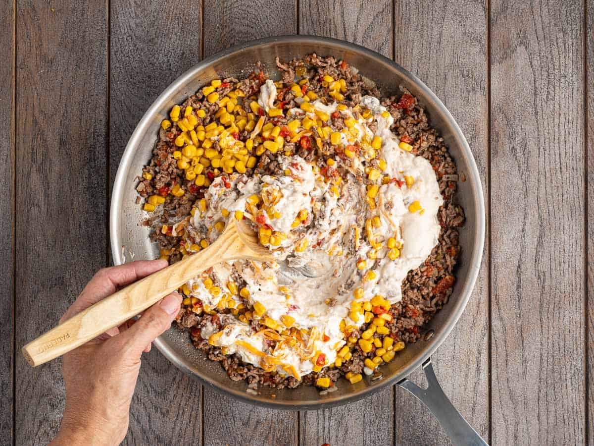 A hand stirs the ground beef, corn, soup and sour cream together in a large skillet on a wooden surface.