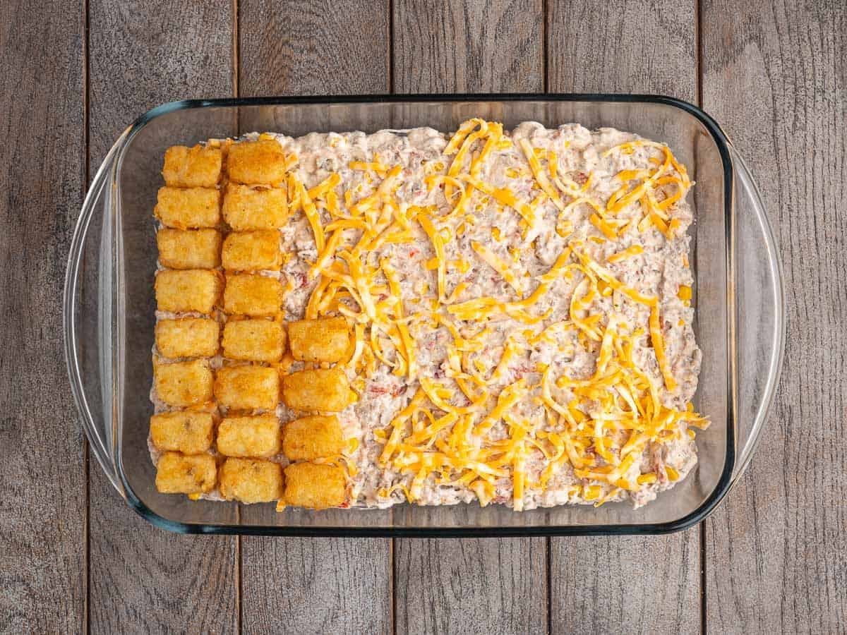 A glass baking dish on a wooden surface holding the cowboy casserole filling topped with shredded cheese, with a row of tater tots laid over one third of the dish.