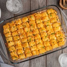 A glass baking dish filled with a golden Cowboy Casserole, topped with melted cheese and chopped green onions, sits on a wooden table next to drinks, plates, and seasonings—capturing all the classic flavors of this hearty Cowboy Casserole Recipe.
