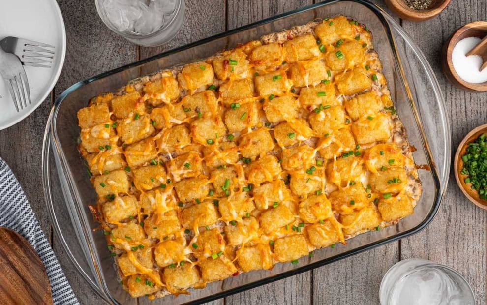 A glass baking dish filled with a golden Cowboy Casserole, topped with melted cheese and chopped green onions, sits on a wooden table next to drinks, plates, and seasonings—capturing all the classic flavors of this hearty Cowboy Casserole Recipe.