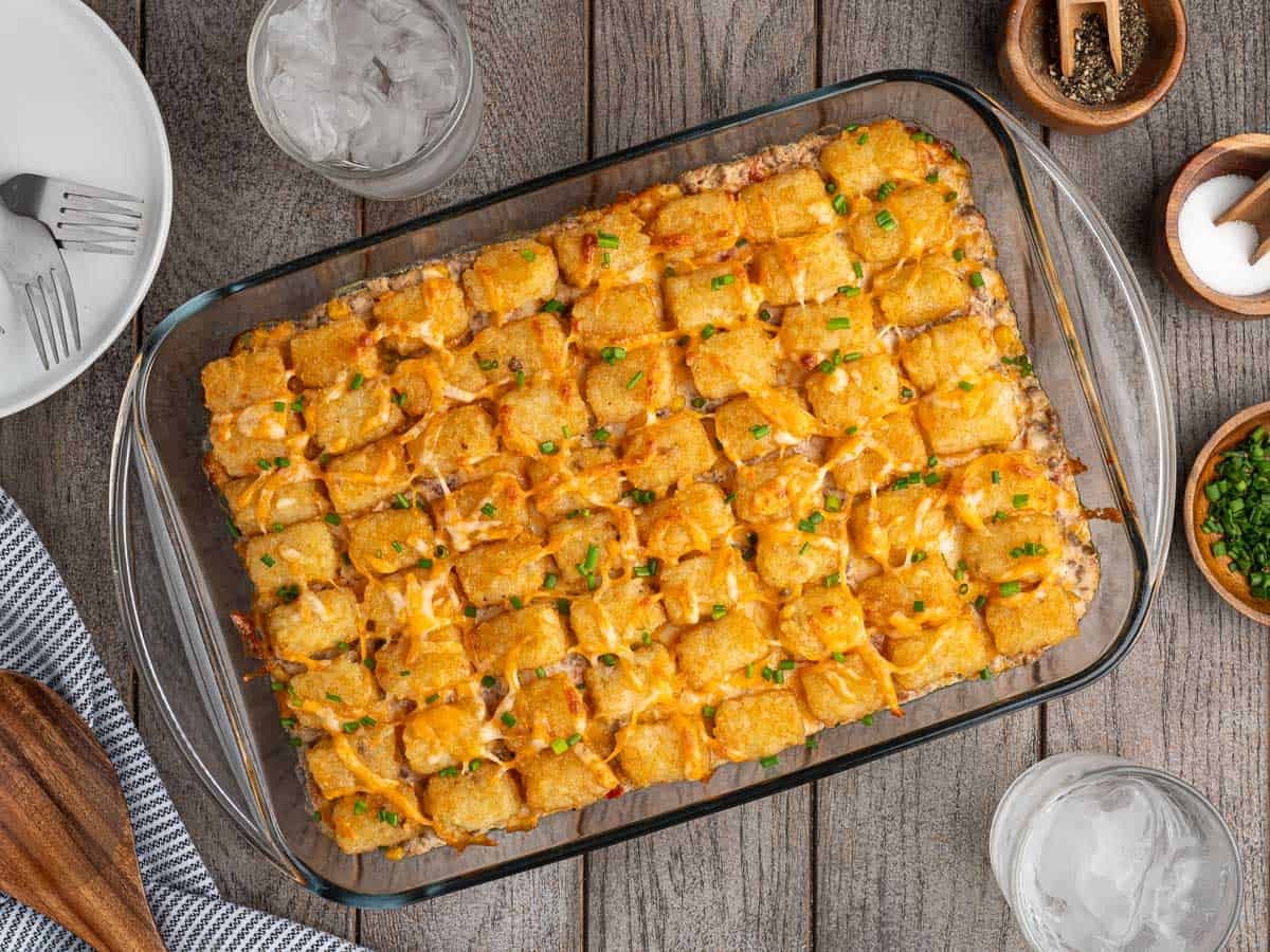 A baked cowboy casserole in a glass dish topped with melted cheese and chopped chives, on a wooden table next to drinks, plates and seasonings.