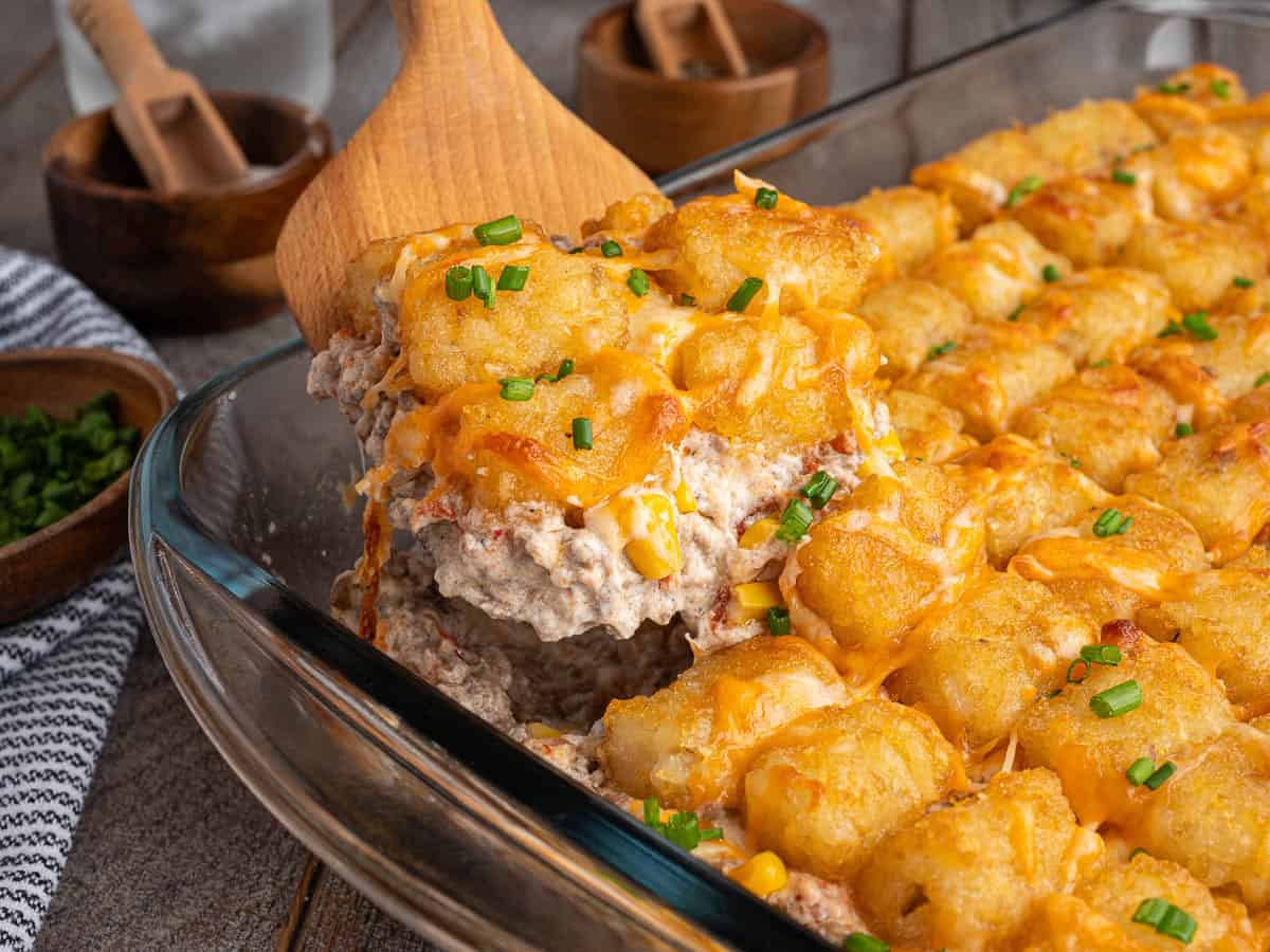 A wooden spoon lifting a serving of cowboy casserole, also known as tater tot hotdish, showing the beef and corn filling under the crisp tater tots and melted cheese.