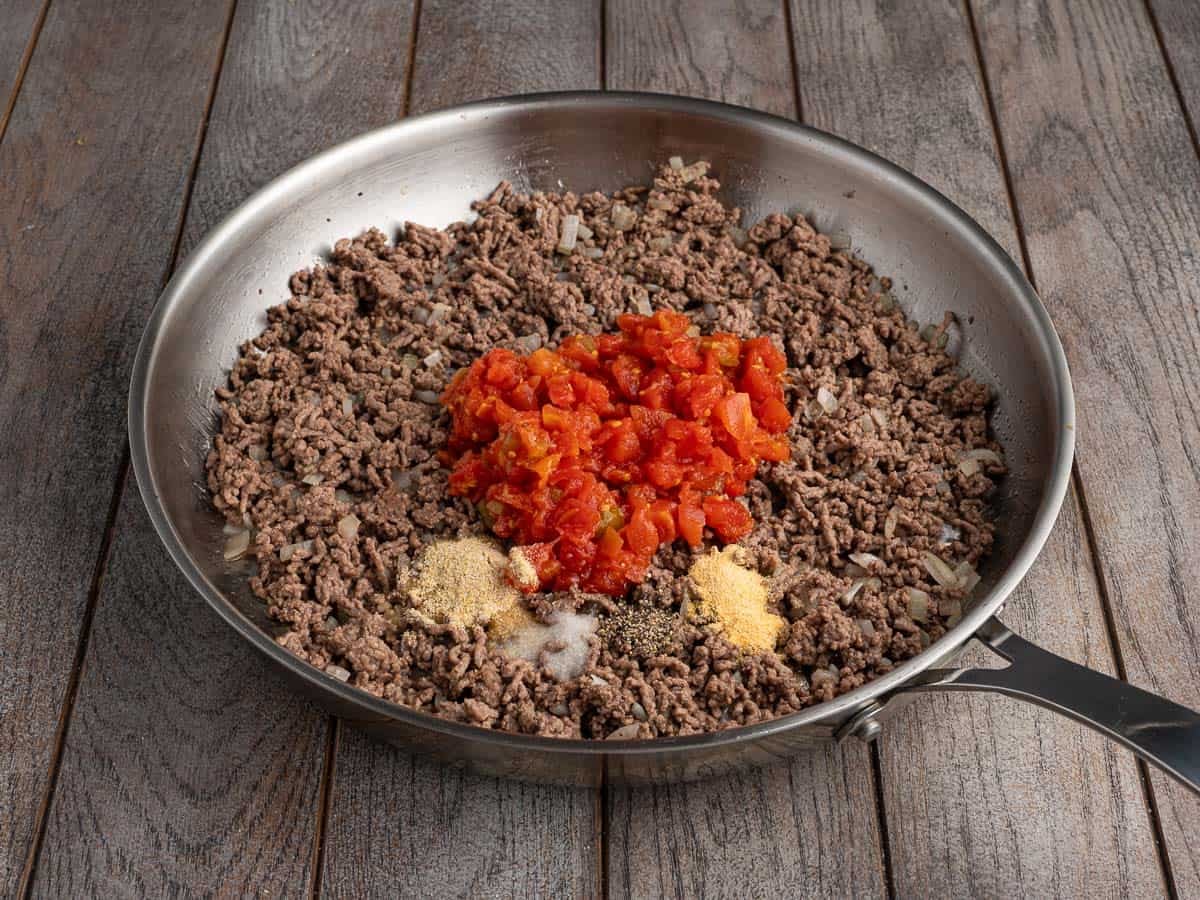 Ground beef, diced onion, Rotel tomatoes and seasonings in a stainless steel skillet on a wooden surface, before cooking.