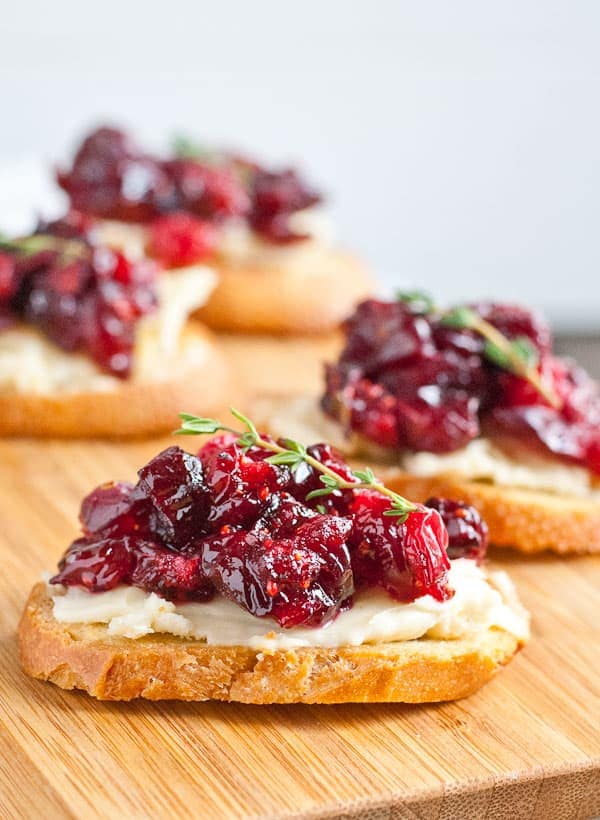 crostini of bread topped with bried and cranberry sauce with a sprig of thyme on top and all sitting on a wood cutting board