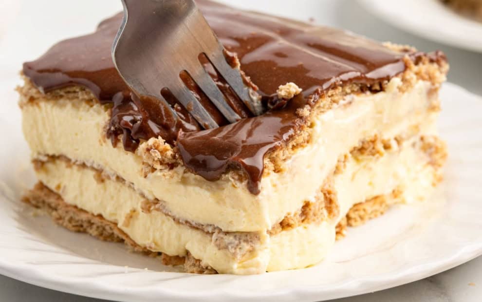 A fork slices into a layered dessert with creamy filling and chocolate topping, served on a white plate—an irresistible Eclair Cake that’s perfect for satisfying your sweet tooth.