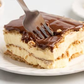 A fork cutting into a slice of layered dessert with creamy filling and chocolate topping on a white plate—this tempting treat is an Eclair Cake, effortlessly capturing all the classic flavors you love in an easy Eclair Cake Recipe.