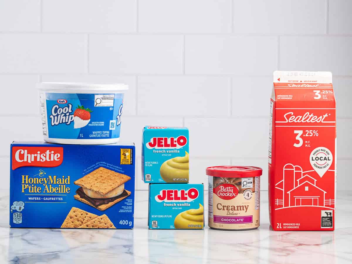 Five grocery items on a marble counter—Cool Whip tub, Honey Maid graham crackers, two Jell-O French vanilla pudding boxes, Betty Crocker chocolate frosting, and a Sealtest milk carton—are neatly arranged in front of a white tile wall, ready to be combined into a delicious Eclair Cake Recipe.