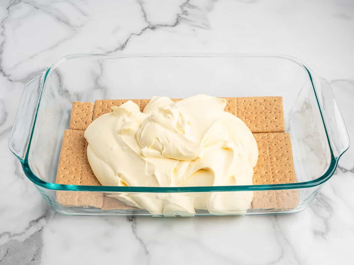 Glass baking dish with a layer of graham crackers on the bottom, topped with a mound of creamy white filling, on a marble countertop—an irresistible start to this classic Eclair Cake Recipe.