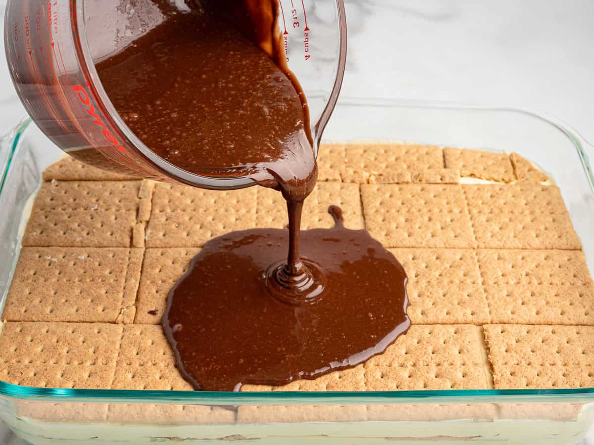 Chocolate sauce is being poured from a measuring cup over a layer of graham crackers in a glass baking dish, likely as part of preparing an Eclair Cake recipe.