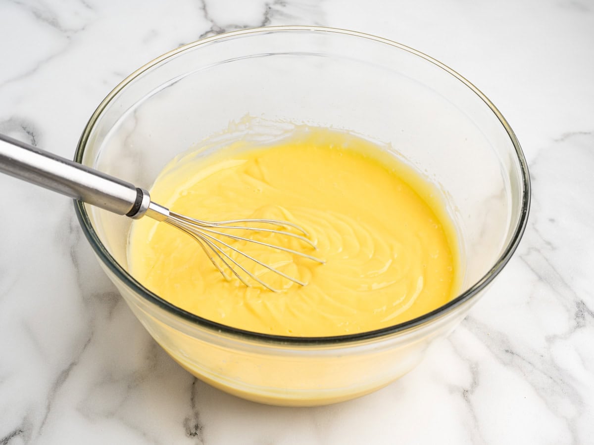 A metal whisk rests in a glass bowl filled with smooth yellow batter on a white marble surface, ready to be transformed into a delicious Eclair Cake.