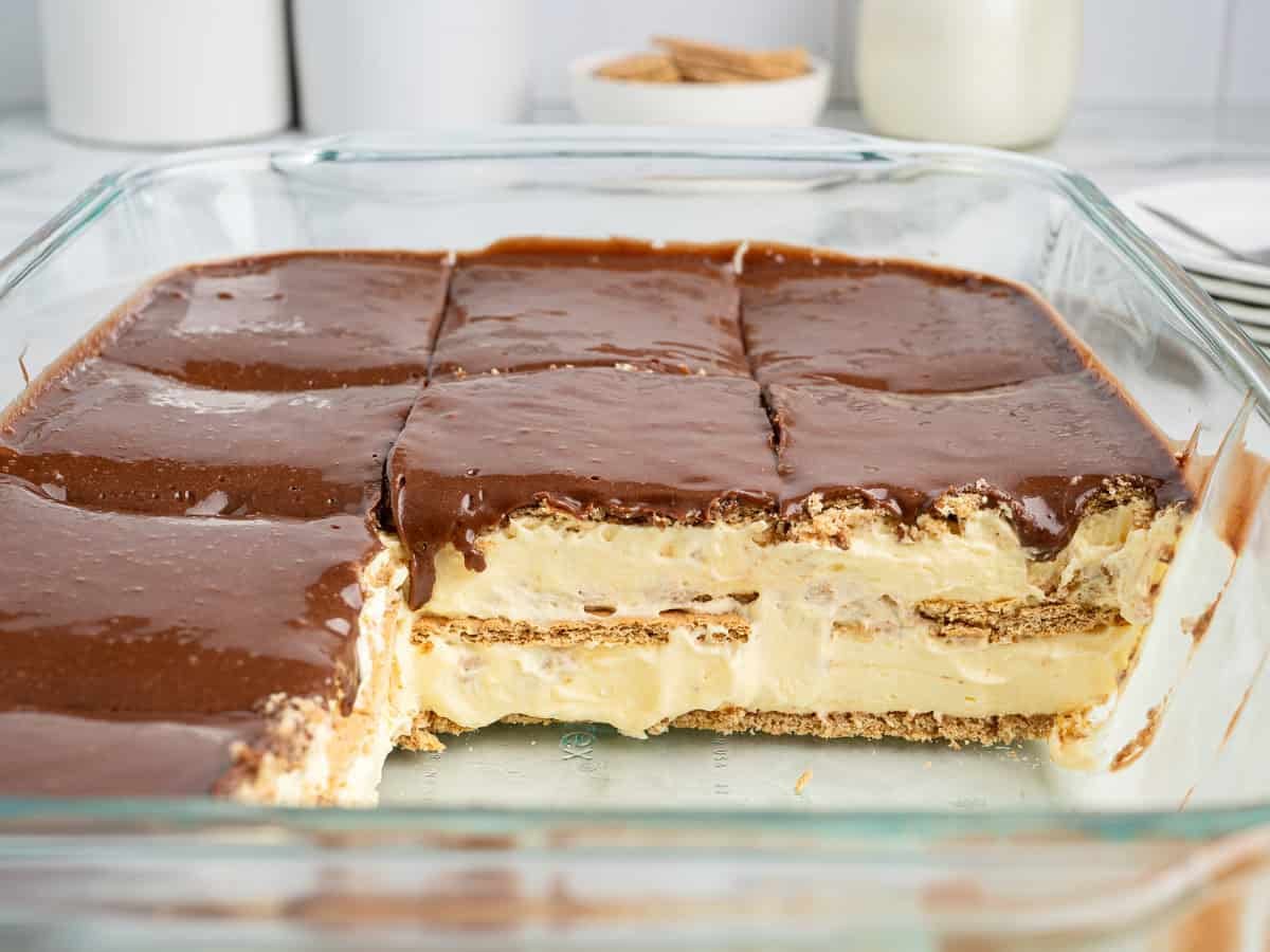 A glass baking dish with an Eclair Cake sliced into squares, showcasing creamy filling and cracker layers beneath a rich chocolate topping. One piece is missing from the dessert, offering a tempting preview of this classic Eclair Cake Recipe. Items are blurred in the background.