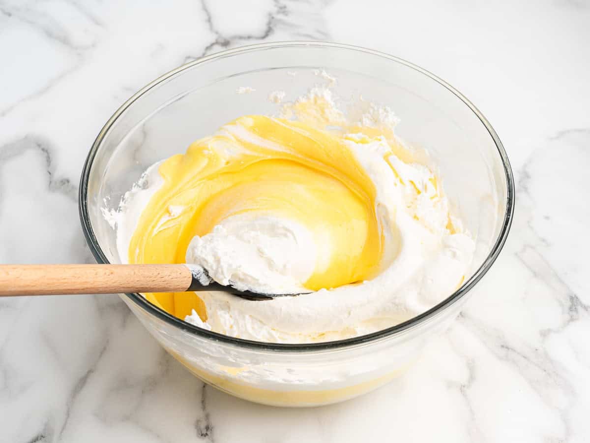A glass bowl filled with whipped cream and yellow cake batter, being gently mixed with a spatula on a marble countertop, creates the perfect base for an irresistible Eclair Cake recipe.