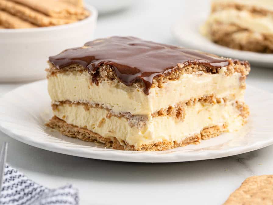 A slice of decadent Eclair Cake sits on a white plate, showcasing its layers of graham crackers, creamy filling, and rich chocolate topping—an irresistible treat from any classic Eclair Cake Recipe.