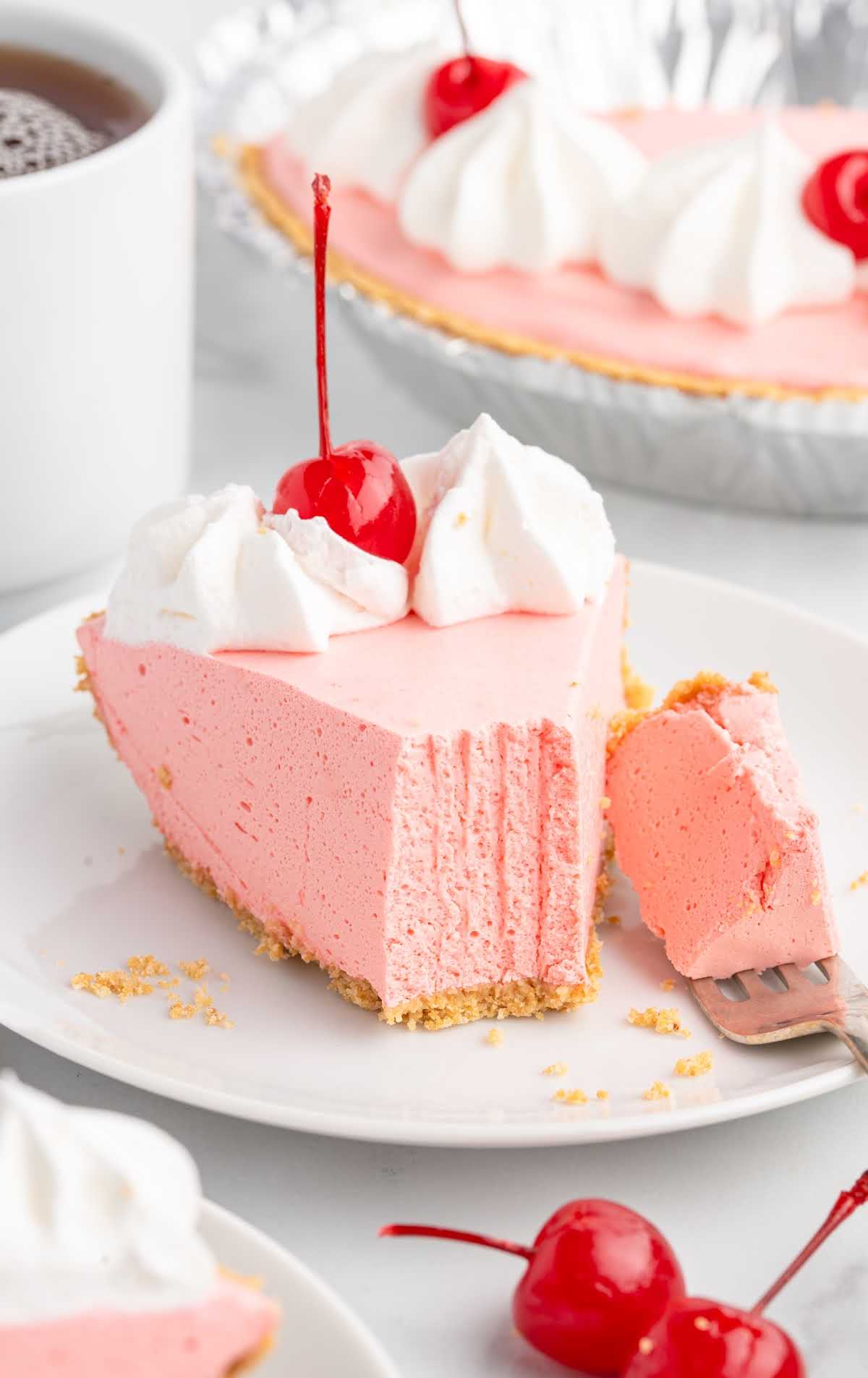Slice of strawberry jello pie on a white plate with a bite taken out, served with whipped cream and a cherry on top.