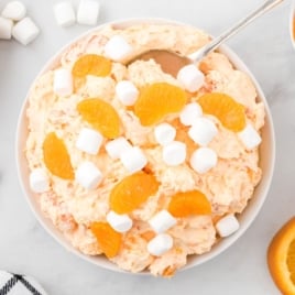 A bowl of Orange Creamsicle Salad topped with mandarin orange slices and mini marshmallows, with extra marshmallows, oranges, and mandarin slices scattered around it on a white surface.