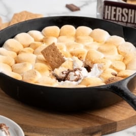A cast iron skillet filled with toasted marshmallows sits on a wooden board. A graham cracker is dipped into the melted S'mores Dip, revealing chocolate underneath. A Hershey&rsquo;s chocolate bar is in the background.