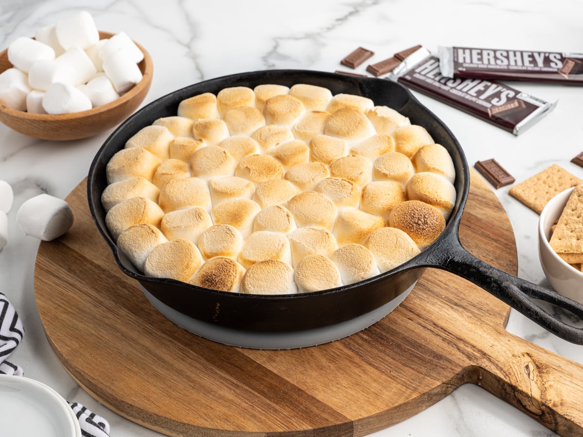 A skillet filled with golden-brown toasted marshmallows sits on a wooden board. Nearby are Hershey&rsquo;s chocolate bars, graham crackers, and a bowl of marshmallows&mdash;perfect for trying out this delicious S'mores Dip Recipe.