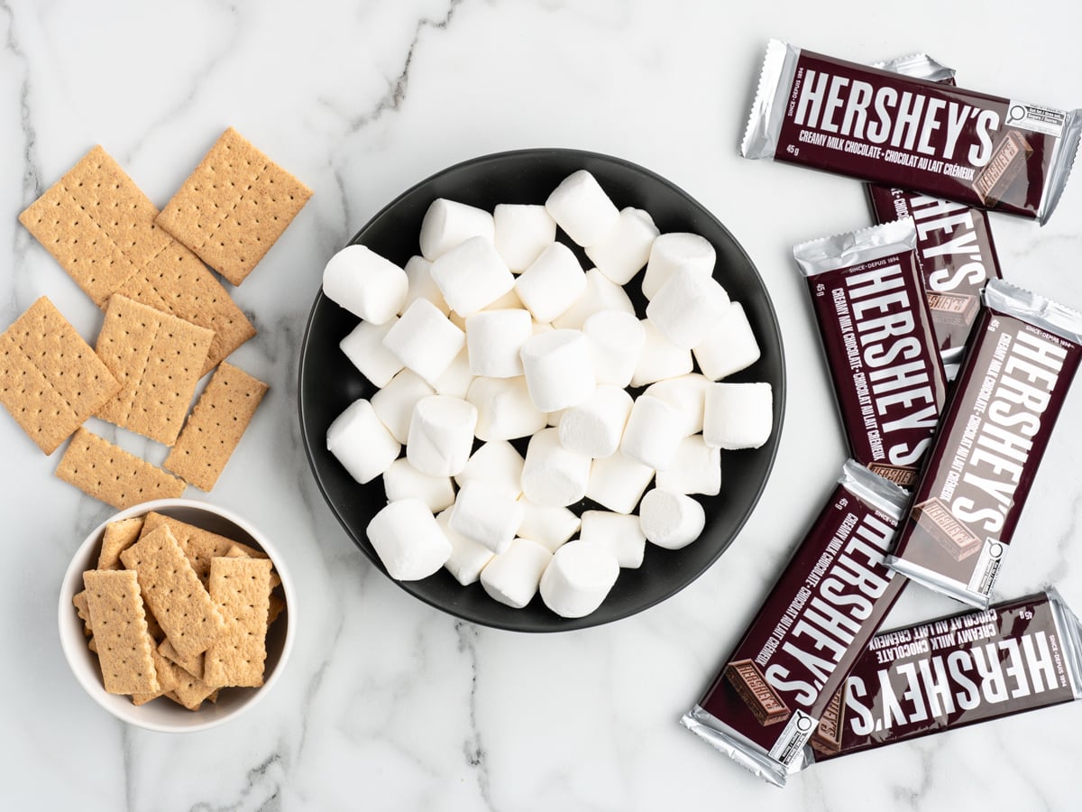 Top-down view of a black bowl filled with marshmallows, Hershey&rsquo;s chocolate bars, and two bowls of graham crackers on a white marble surface. Perfect ingredients arranged separately for making a classic S'mores Dip recipe.