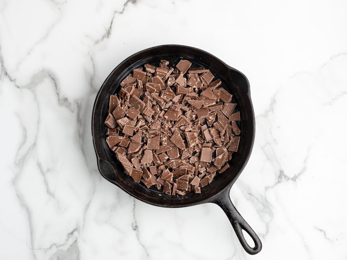 A black cast iron skillet filled with chopped milk chocolate pieces sits on a white marble surface, ready to be transformed into a delicious S'mores Dip.
