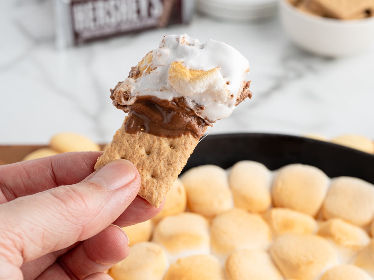 A hand holds a graham cracker topped with melted chocolate and gooey toasted marshmallow, with a skillet of golden S'mores Dip in the background. Perfect for sharing, this S'mores Dip Recipe brings campfire flavors indoors.