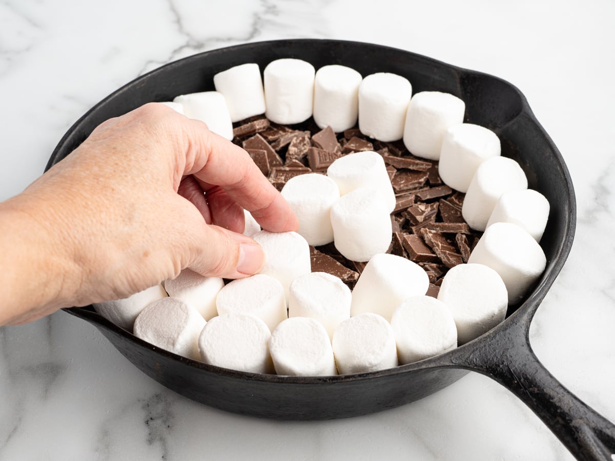 A hand arranges marshmallows in a circle over chopped chocolate pieces in a black cast iron skillet, preparing a delicious S'mores Dip on a white marble surface.