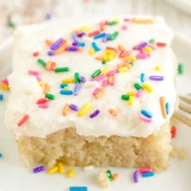 Close-up of a frosted vanilla crazy cake slice with colorful sprinkles on a white plate with a fork.