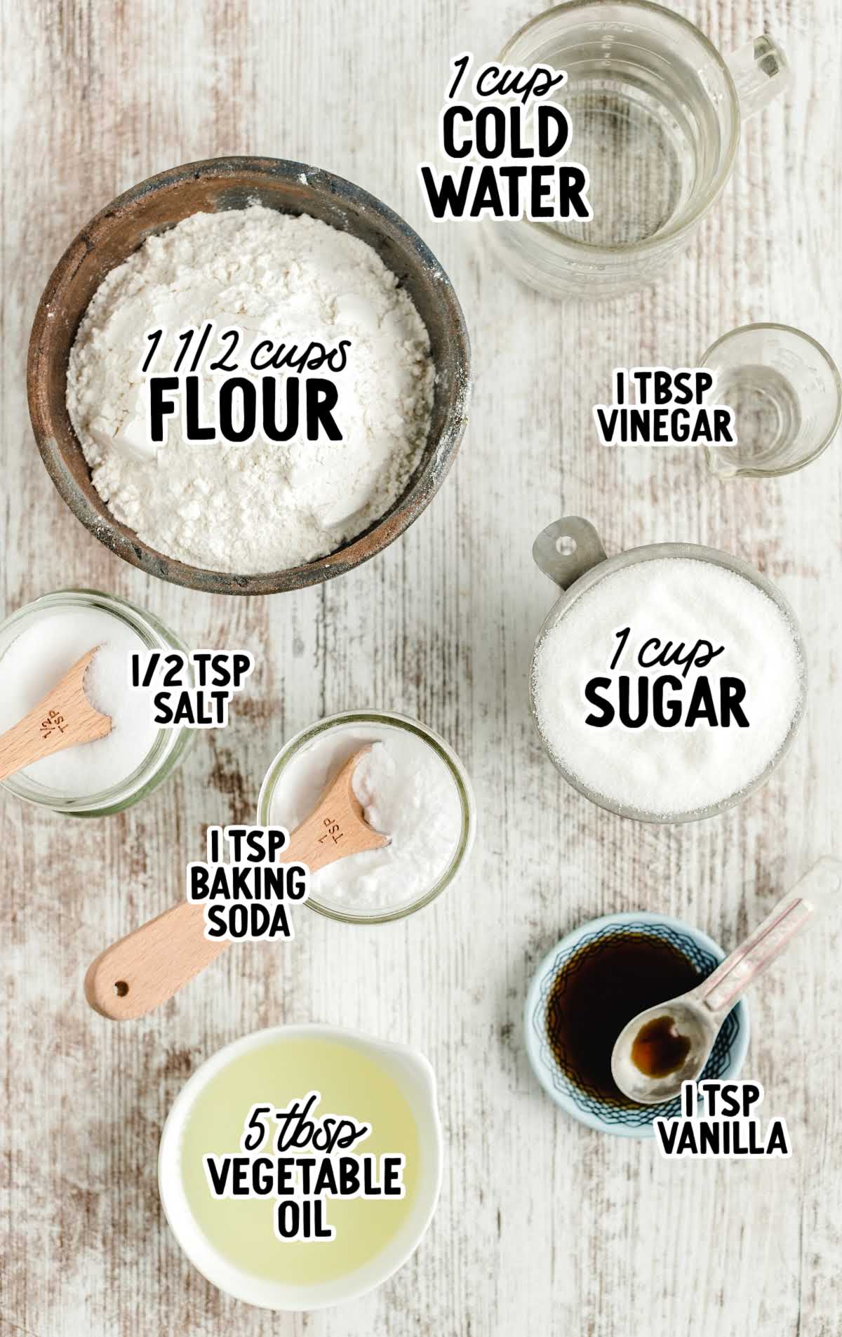 Flat lay of vanilla crazy cake ingredients including flour, sugar, vinegar, vanilla, baking soda, salt, oil, and water with labeled text.