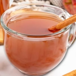 Glass mugs of warm apple cider garnished with cinnamon sticks and apple slices.