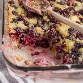 close up shot of blackberry cobbler in a baking dish with a wooden spoon and a serving missing