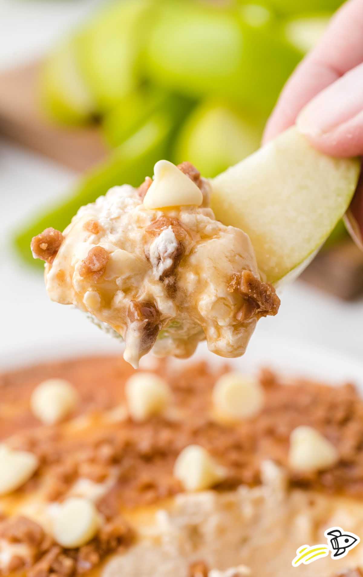 close up shot a slice of apple dipped with caramel apple dip topped with caramel, toffee, and white chocolate