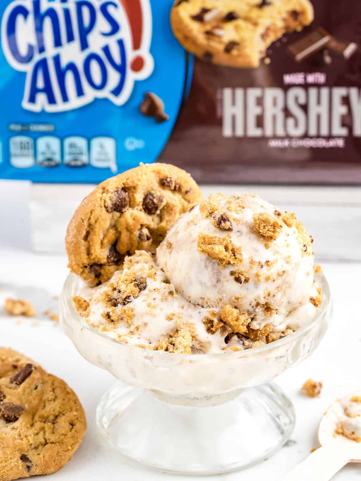 A glass bowl of vanilla ice cream topped with crumbled chocolate chip cookies. Two whole cookies are on the edge. Chips Ahoy and Hersheys cookie packages are visible in the background, making this an irresistible No-Churn Chips Ahoy Ice Cream treat.