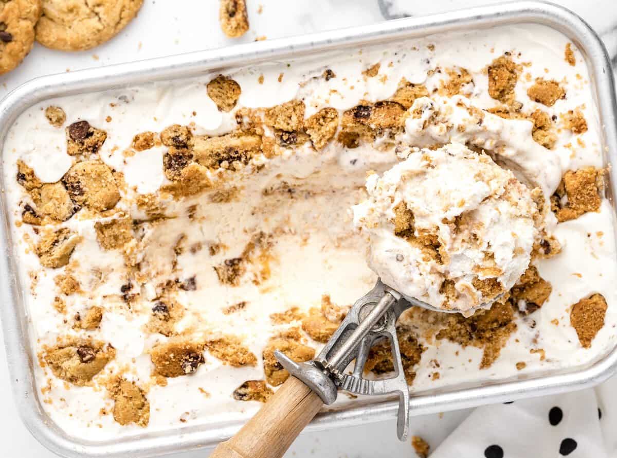 A metal tray of No-Churn Chips Ahoy Ice Cream with chunks of chocolate chip cookies, partially scooped with an ice cream scooper. Pieces of cookies are scattered on top, and a white cloth with black dots is nearby.