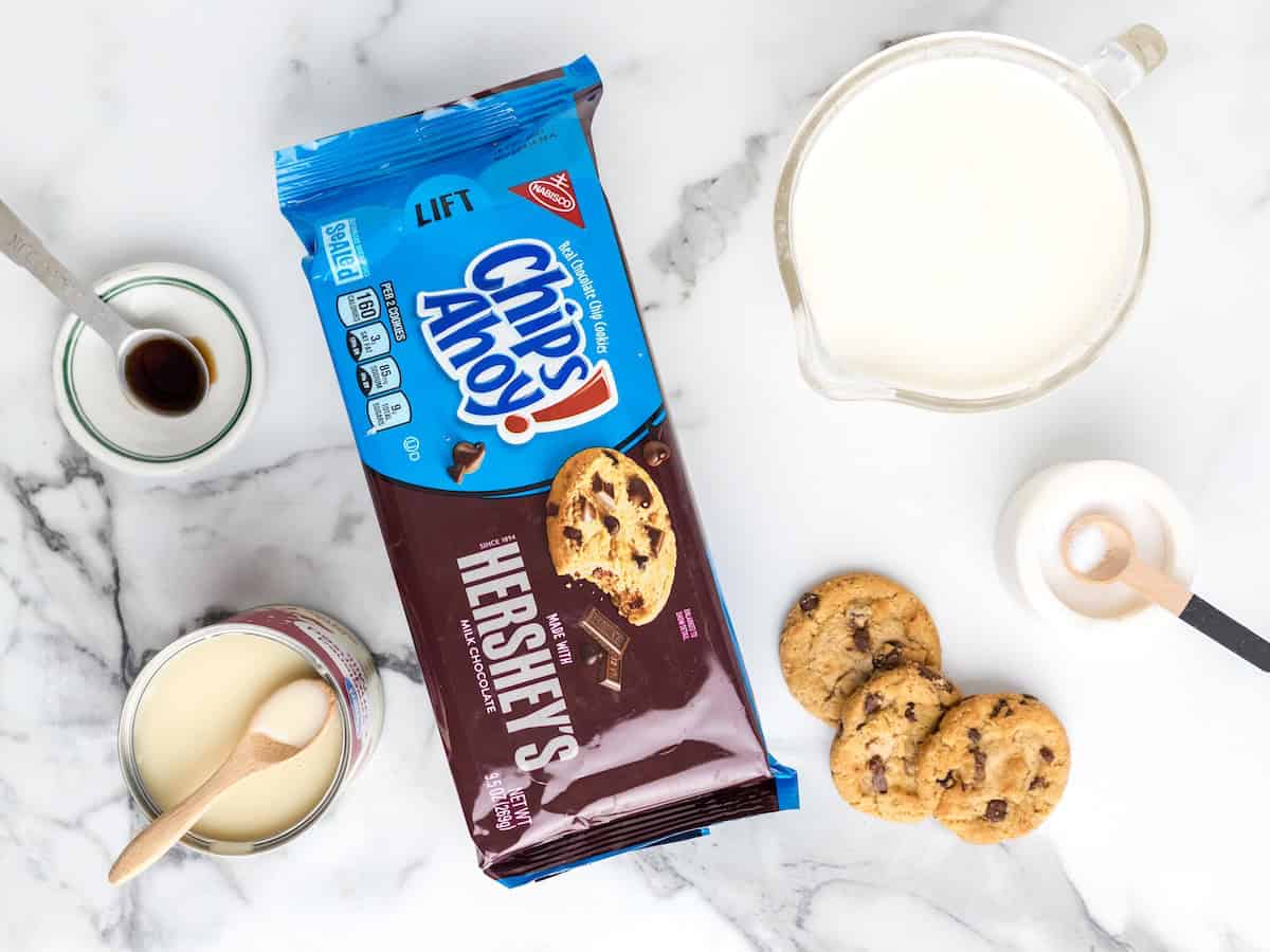 An open package of Chips Ahoy! Hershey’s cookies, a glass jug of milk, a can of condensed milk, and a small bowl of vanilla and sugar sit on a marble surface beside chocolate chip cookies—everything you need to whip up delicious No-Churn Chips Ahoy Ice Cream at home.