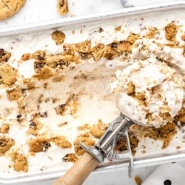 A metal tray of No-Churn Chips Ahoy Ice Cream with chunks of chocolate chip cookies, partially scooped out. An ice cream scoop holds a serving, and crunchy cookie pieces are scattered on top and nearby.