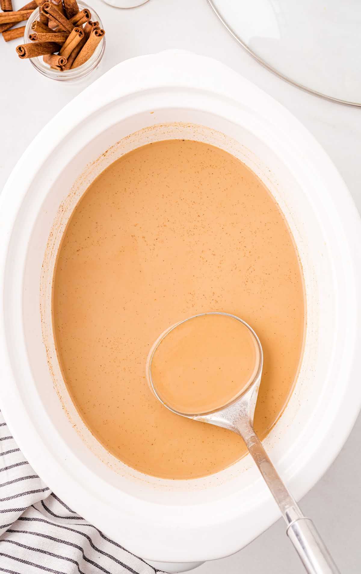 Slow cooker of pumpkin spice latte with ladle surrounded by cinnamon sticks.